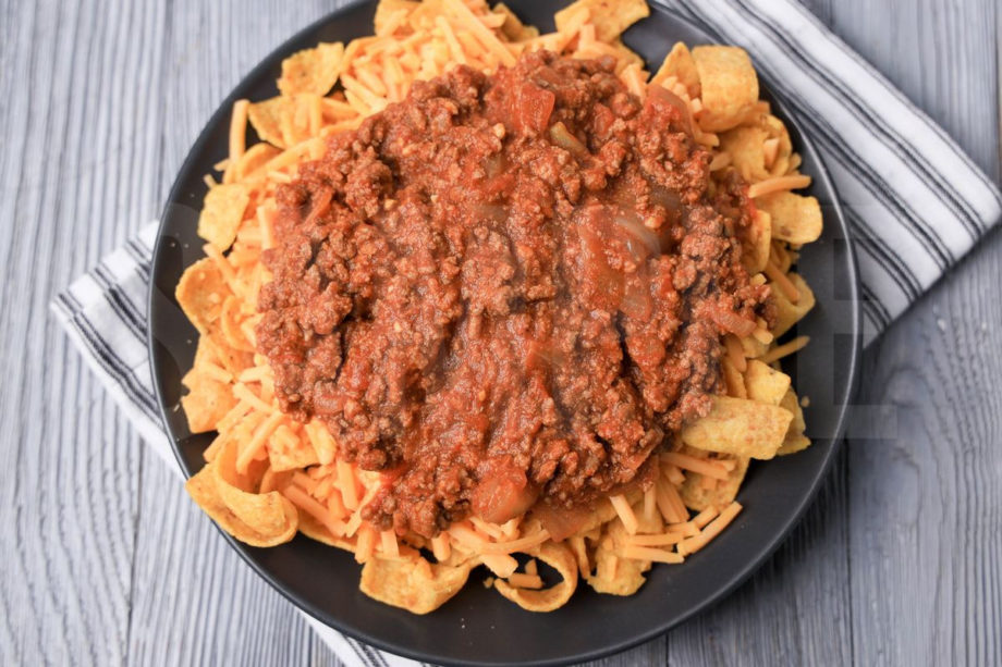 The Frito Pie comes on a gray plate with a white striped napkin on a gray wood backdrop.