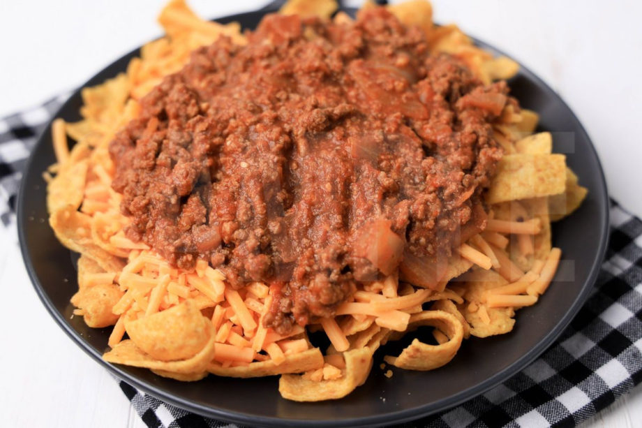 The Frito Pie comes on a gray plate with a plaid napkin on a white wood backdrop.