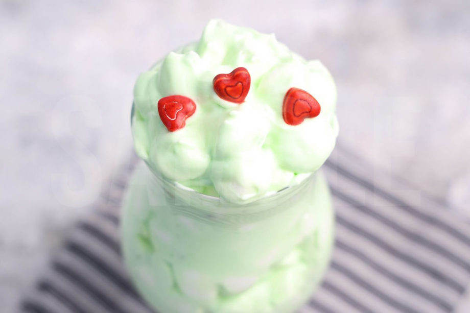 The Grinch Fluff comes in a clear glass with a gray striped napkin on a marble wood backdrop.