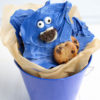 The Cookie Monster Bark comes in a blue pail with parchment paper with a blue and white striped cloth on a white wood backdrop.