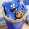 The Cookie Monster Bark comes in a blue pail with parchment paper with a blue and white striped cloth on a white wood backdrop.