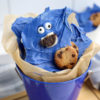 The Cookie Monster Bark comes in a blue pail with parchment paper with a blue and white striped cloth on a white wood backdrop.