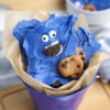 The Cookie Monster Bark comes in a blue pail with parchment paper with a blue and white striped cloth on a white wood backdrop.