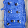 The Cookie Monster Bark comes in a blue pail with parchment paper with a blue and white striped cloth on a white wood backdrop.