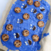 The Cookie Monster Bark comes in a blue pail with parchment paper with a blue and white striped cloth on a white wood backdrop.