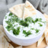 The Tzatziki Dip comes in a white bowl on a wood cutting board with a olive green cloth on a white wood backdrop.