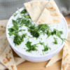 The Tzatziki Dip comes in a white bowl on a wood cutting board with a olive green cloth on a white wood backdrop.