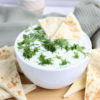 The Tzatziki Dip comes in a white bowl on a wood cutting board with a olive green cloth on a white wood backdrop.