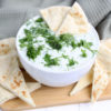 The Tzatziki Dip comes in a white bowl on a wood cutting board with a olive green cloth on a white wood backdrop.