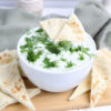 The Tzatziki Dip comes in a white bowl on a wood cutting board with a olive green cloth on a white wood backdrop.