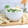The Tzatziki Dip comes in a white bowl on a wood cutting board with a olive green cloth on a white wood backdrop.