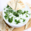 The Tzatziki Dip comes in a white bowl on a wood cutting board with a olive green cloth on a white wood backdrop.