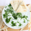 The Tzatziki Dip comes in a white bowl on a wood cutting board with a olive green cloth on a white wood backdrop.