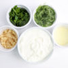 The Tzatziki Dip comes in a white bowl on a wood cutting board with a olive green cloth on a white wood backdrop.