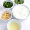 The Tzatziki Dip comes in a white bowl on a wood cutting board with a olive green cloth on a white wood backdrop.