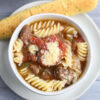 The Slow Cooker Beef Parmesan Soup comes on a white bowl with gray wood backdrop.