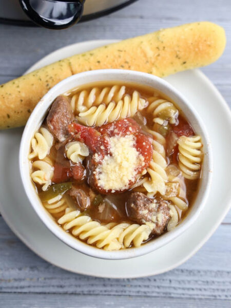 The Slow Cooker Beef Parmesan Soup comes on a white bowl with gray wood backdrop.