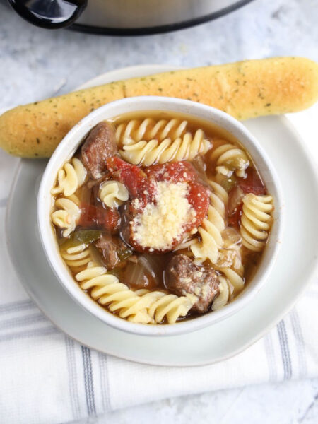 The Slow Cooker Beef Parmesan Soup comes on a white bowl with marble wood backdrop.