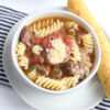 The Slow Cooker Beef Parmesan Soup comes on a white bowl with white wood backdrop.