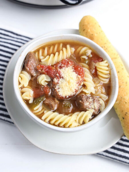 The Slow Cooker Beef Parmesan Soup comes on a white bowl with white wood backdrop.